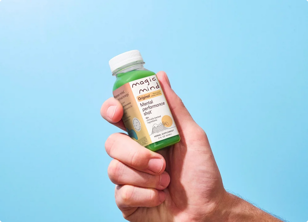 Hand holding a Magic Mind Original Mental Performance Shot bottle against a blue background.