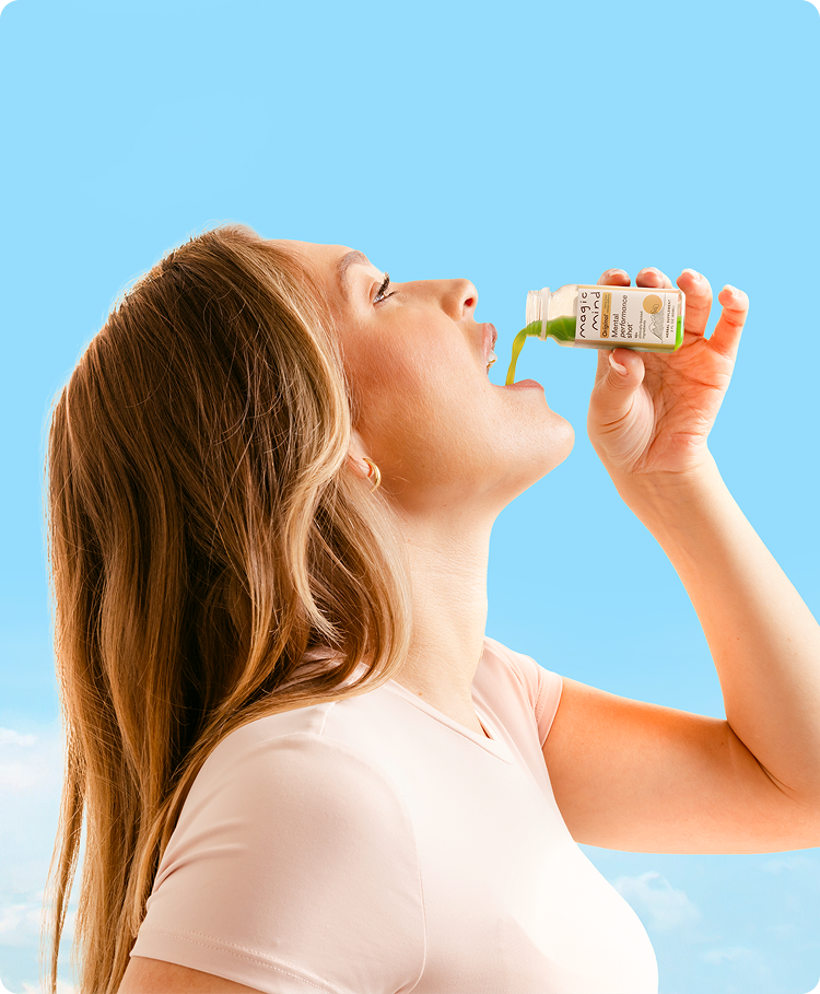 Woman drinking an Original Magic Mind mental performance shot directly from the bottle, head tilted back against a blue sky background.