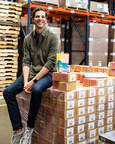 James Beshara, founder of Magic Mind, sitting on a pallet of boxes in a warehouse filled with inventory, smiling at the camera.