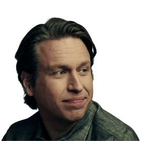 Pete Holmes; headshot of a man with light brown hair wearing a dark jacket, with a slight smile against a transparent background