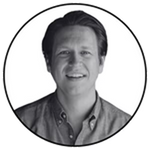 Black and white circular headshot of Pete Holmes smiling, wearing a button-down shirt.