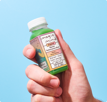 Hand holding a Magic Mind Original Mental Performance Shot bottle against a light blue background