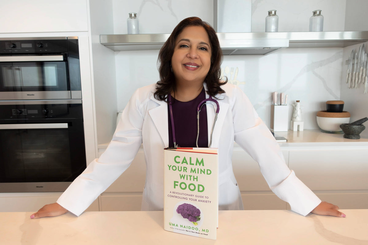 Uma Naidoo in a white coat holding the book 'Calm Your Mind with Food' by Uma Naidoo, MD, in a kitchen.