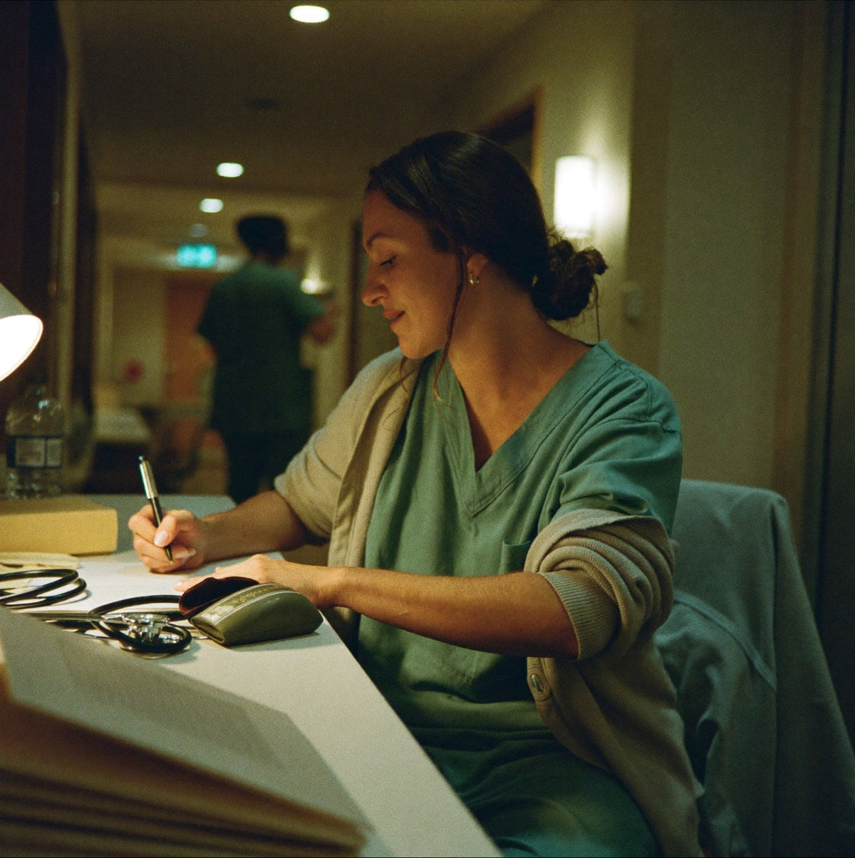 Healthcare worker in scrubs writing notes at a desk during a night shift.