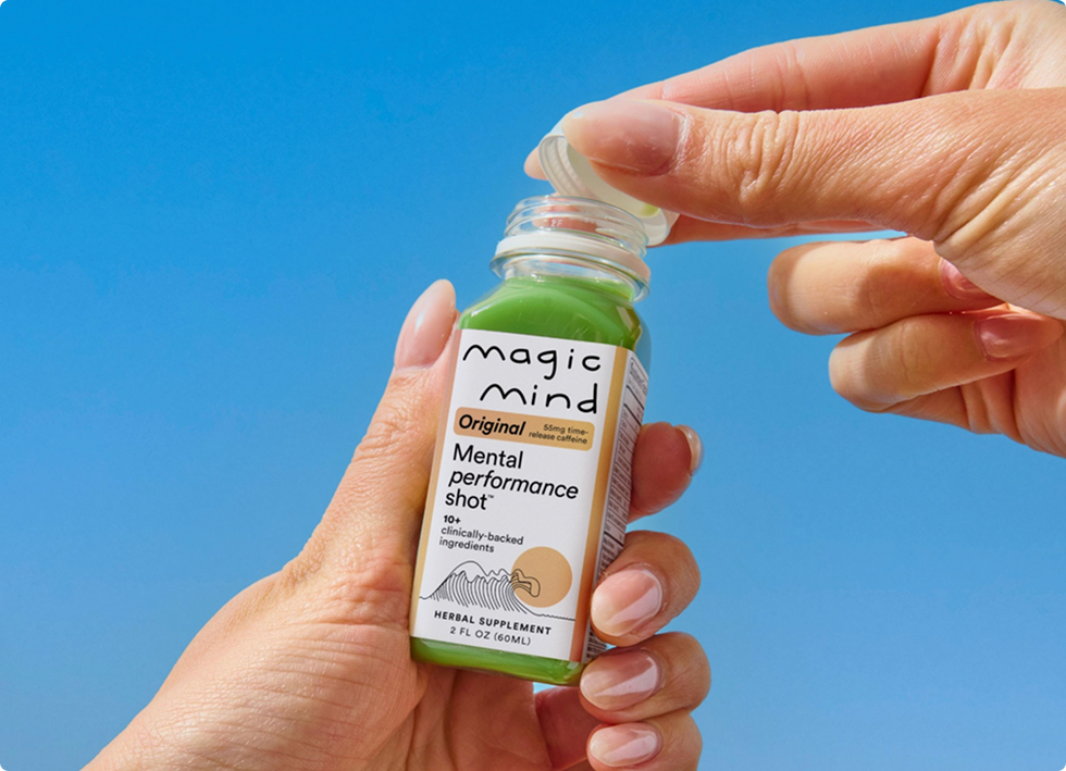Hands opening a Magic Mind Original Mental Performance Shot bottle against a blue sky.