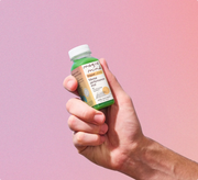 Hand holding a Magic Mind Original Mental Performance Shot bottle against a pink gradient background.
